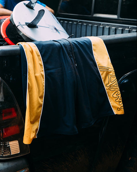 Black jacket with yellow accents draped over a car's rear, with a surfboard in the background.