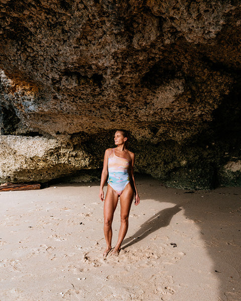 Woman in a swimsuit standing on a sandy beach with large rocks and cave entrance in the background