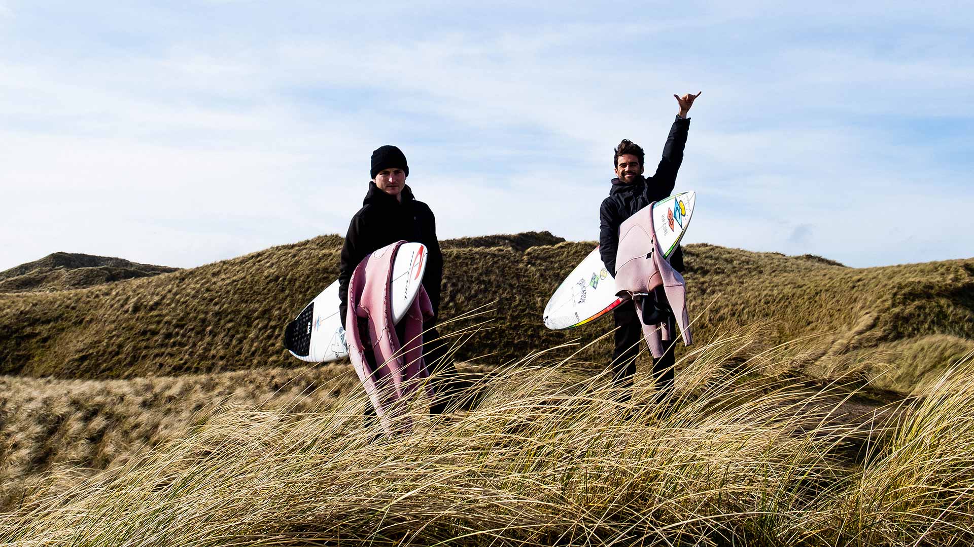 Mason Ho and friend on the windy hills of Scotland holding their boards and Rip Curl wetsuits