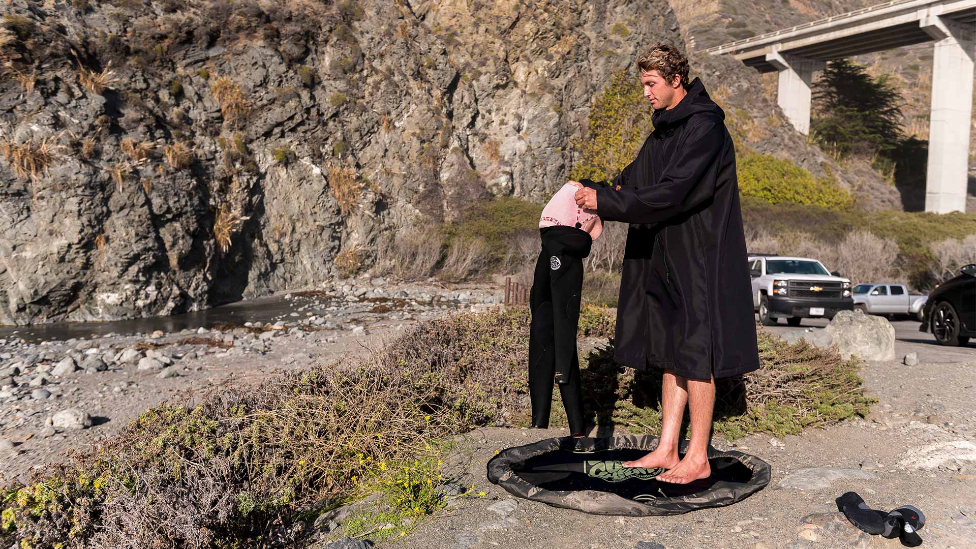 Conner Coffin holding his surfboard and walking along the cobblestones at the water’s edge wearing his hooded Rip Curl Flashbomb Wetsuit and booties.