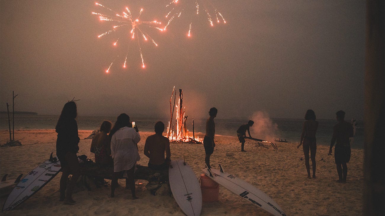 Rip Curl Women standing around a fire on the beach with fireworks in the sky