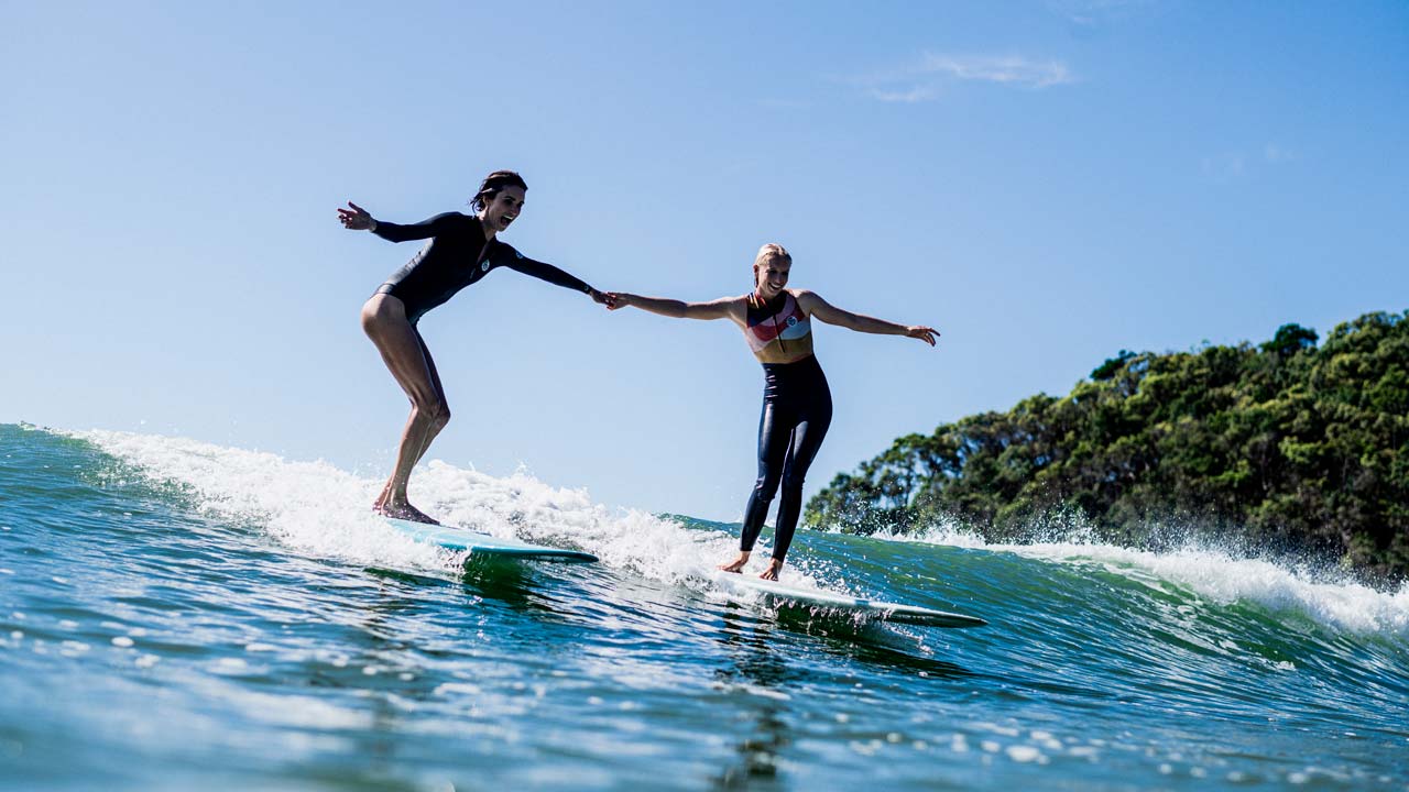 Victoria Vergara and friend holding hands surfing longboards next to each other.