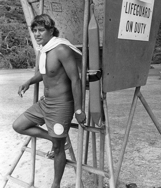 Black and white image of Eddie Aikau standing against a lifesaving tower