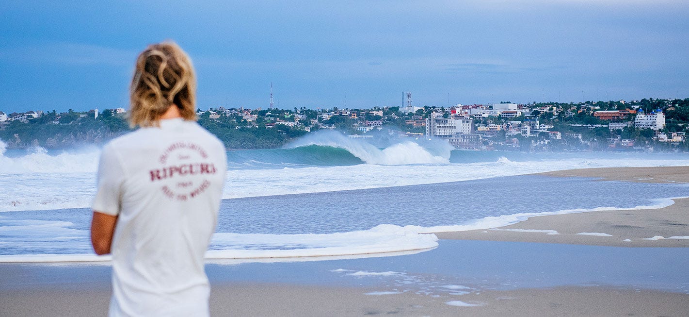 Man in white Rip Curl shirt, staring across the waves at a town on the other side of the bay